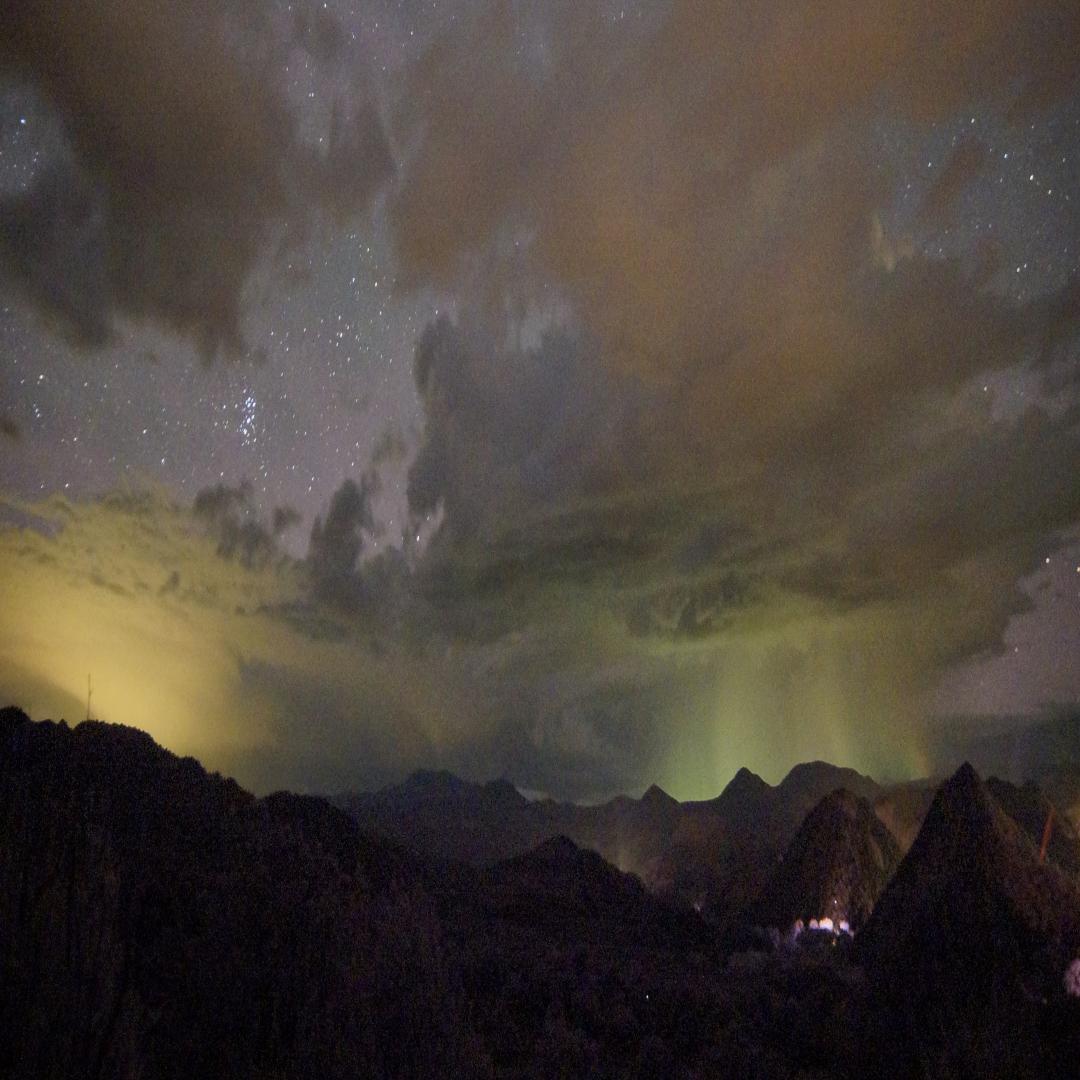 A mountainous scene is shown at night, with a small, glowing city in the lower right-hand corner. The mountains are dwarfed by an enormous sky, peppered with patchy clouds and swaths of stars. The sky has an unnatural glow (literally), with fields of yellow-green and orangish light, indicating light pollution.