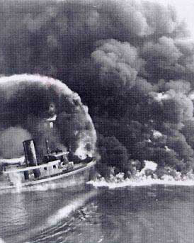 A firefighter sprays water from a bridge onto the burning Cuyahoga River in this historical photo, in black and white..