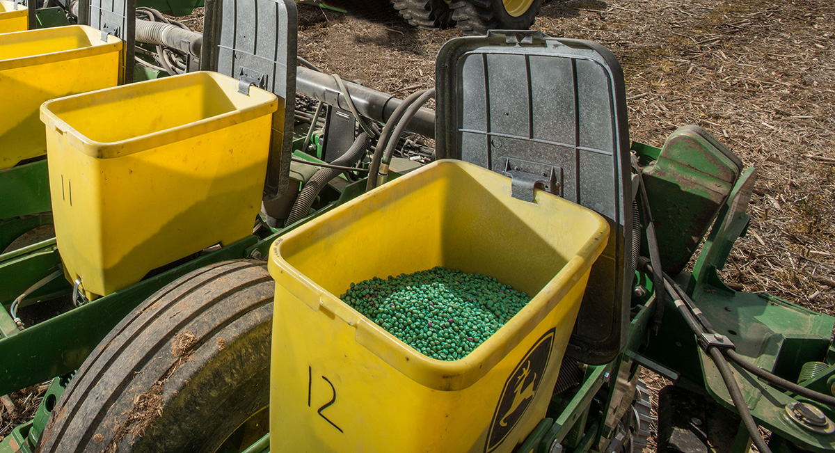 A series of seed-dispersing machines in a row. The main container is open, showing a pile of bright green, neonic-coated seeds.
