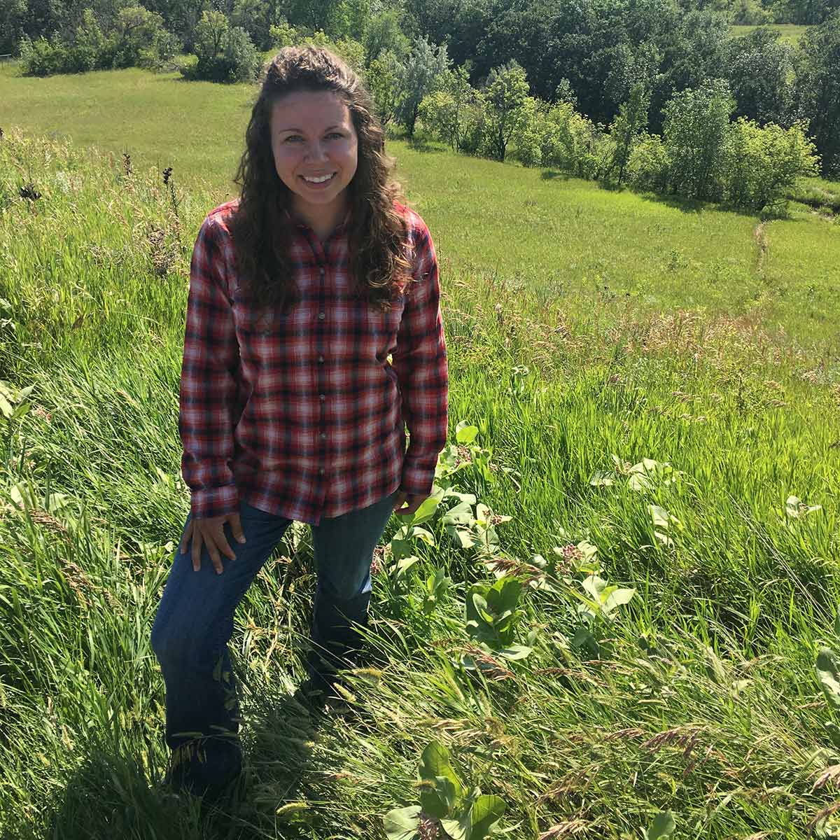 A white woman wearing a flannel shirt, standing in a grassland and smiling at the camera