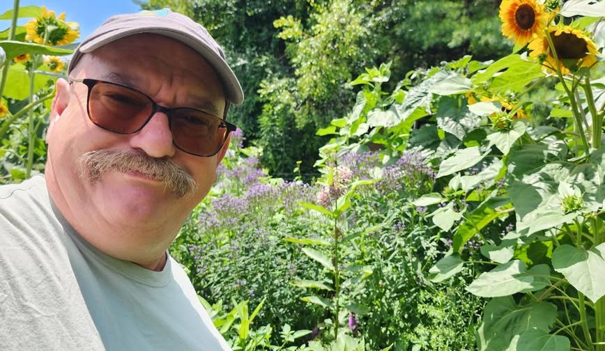 Michael McIntyre smiling in front of tall flowering plants like sunflowers