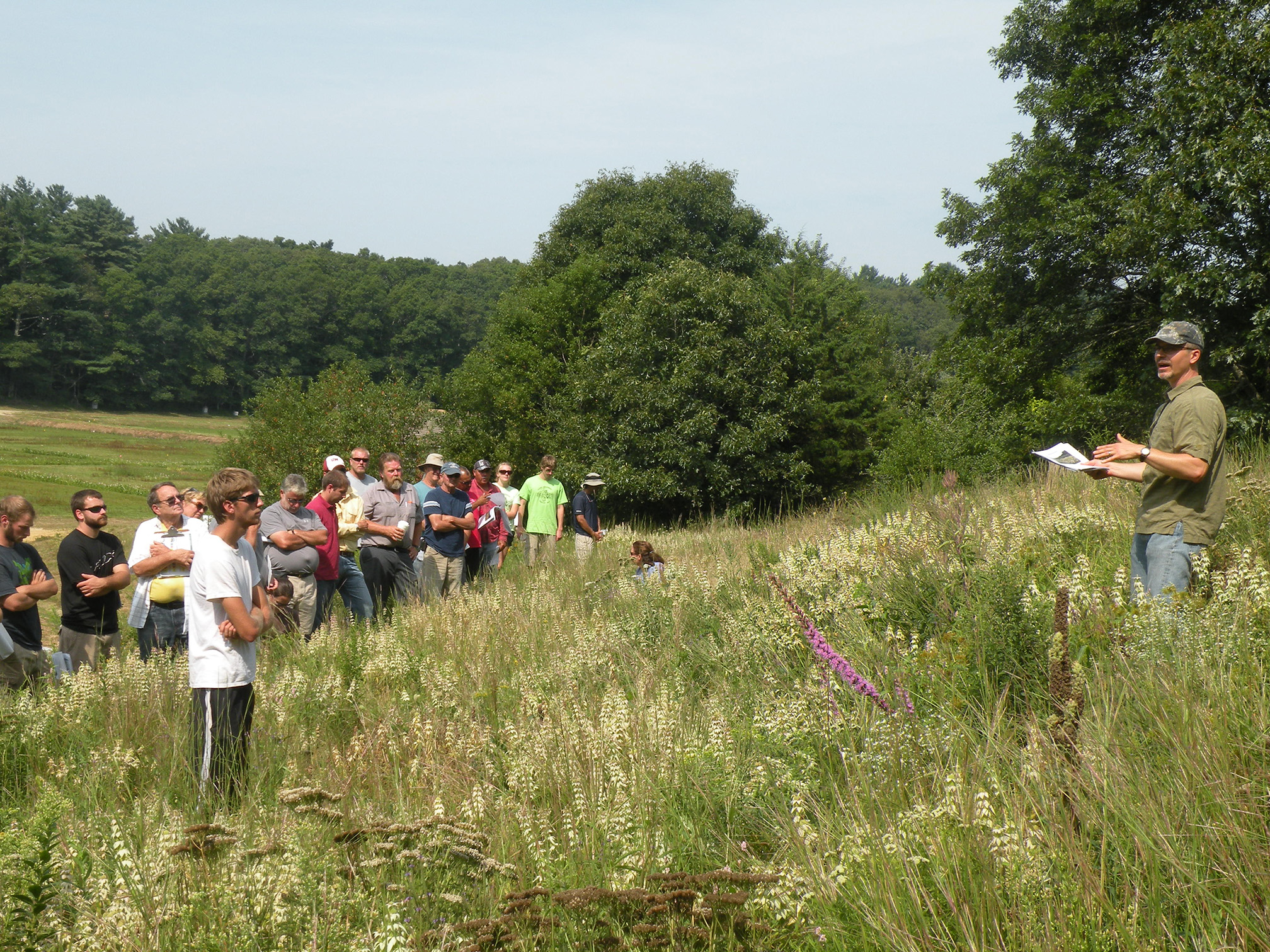 A group of men and women of various ages stand listening to a man wearing a green shirt and gray cap. The ground between them is full of long grass and white flowers.