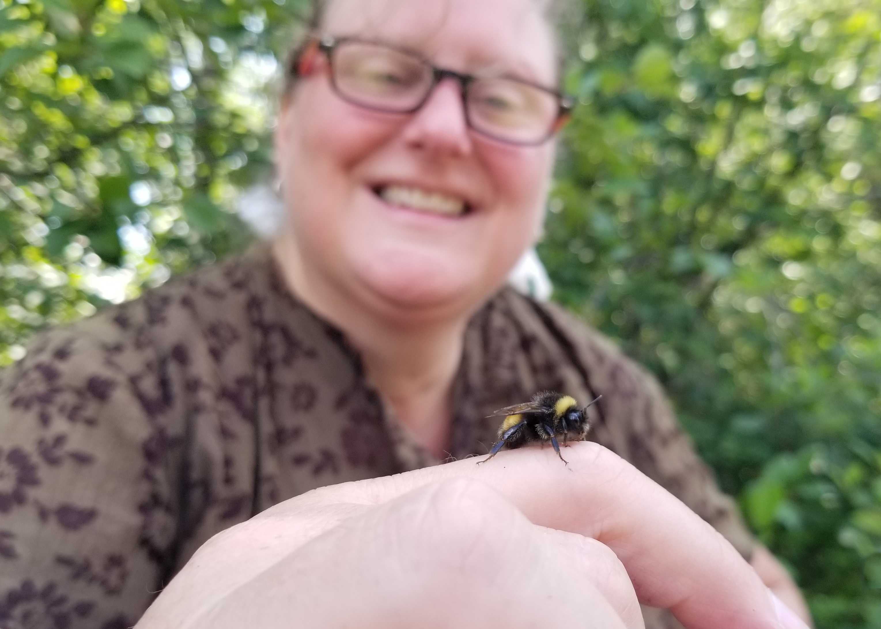 A woman smiles while looking at a fuzzy, black, and yellow bumble bee on her finger.
