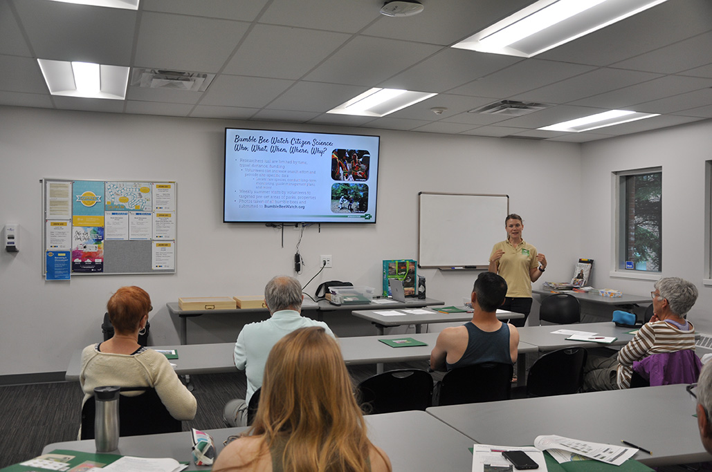 People sit at long tables in a classroom as an instructor leads a discussion.