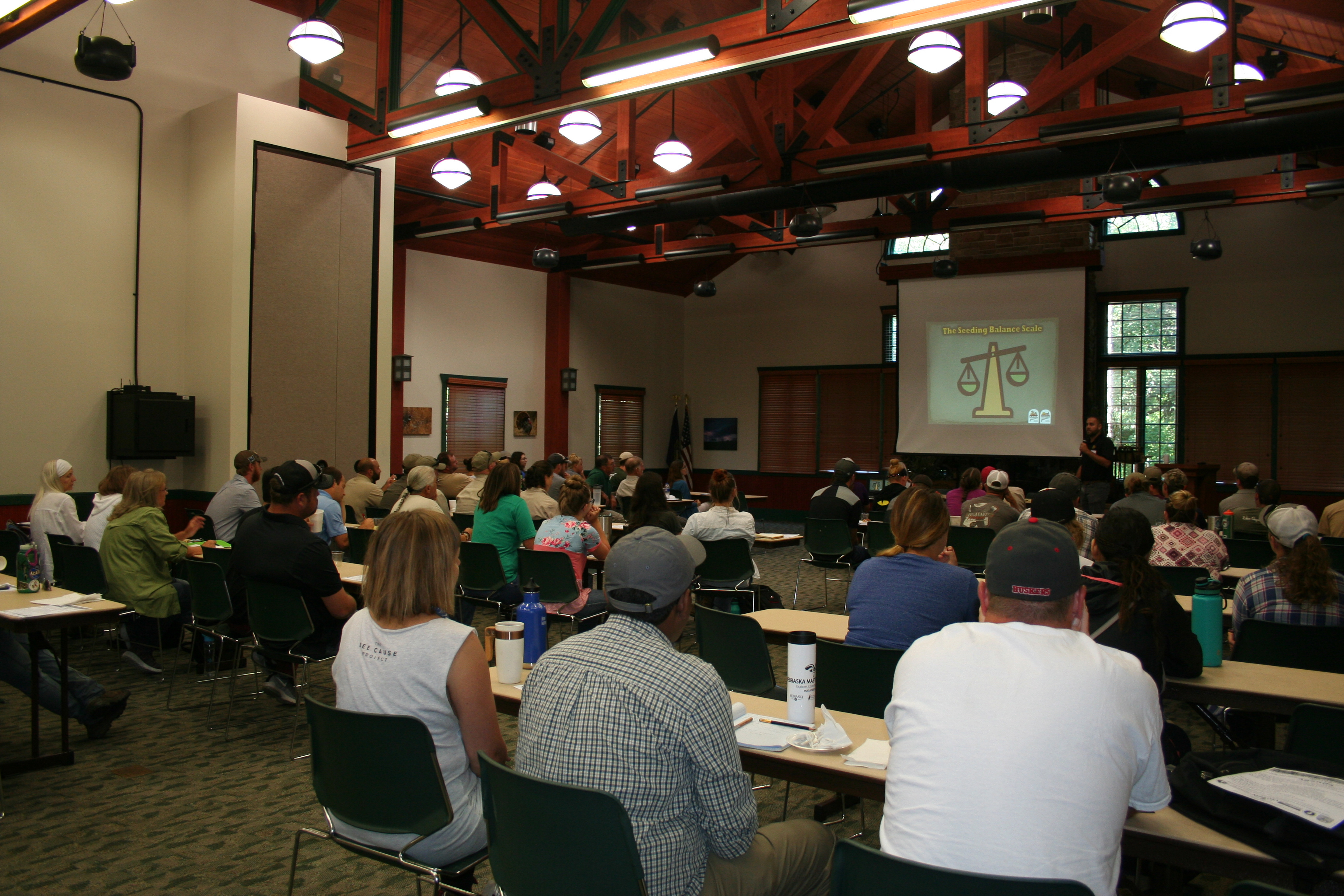 Many people are seated in a room. Their backs are to the camera. They are looking up at a screen on which a Powerpoint presentation is being projected.