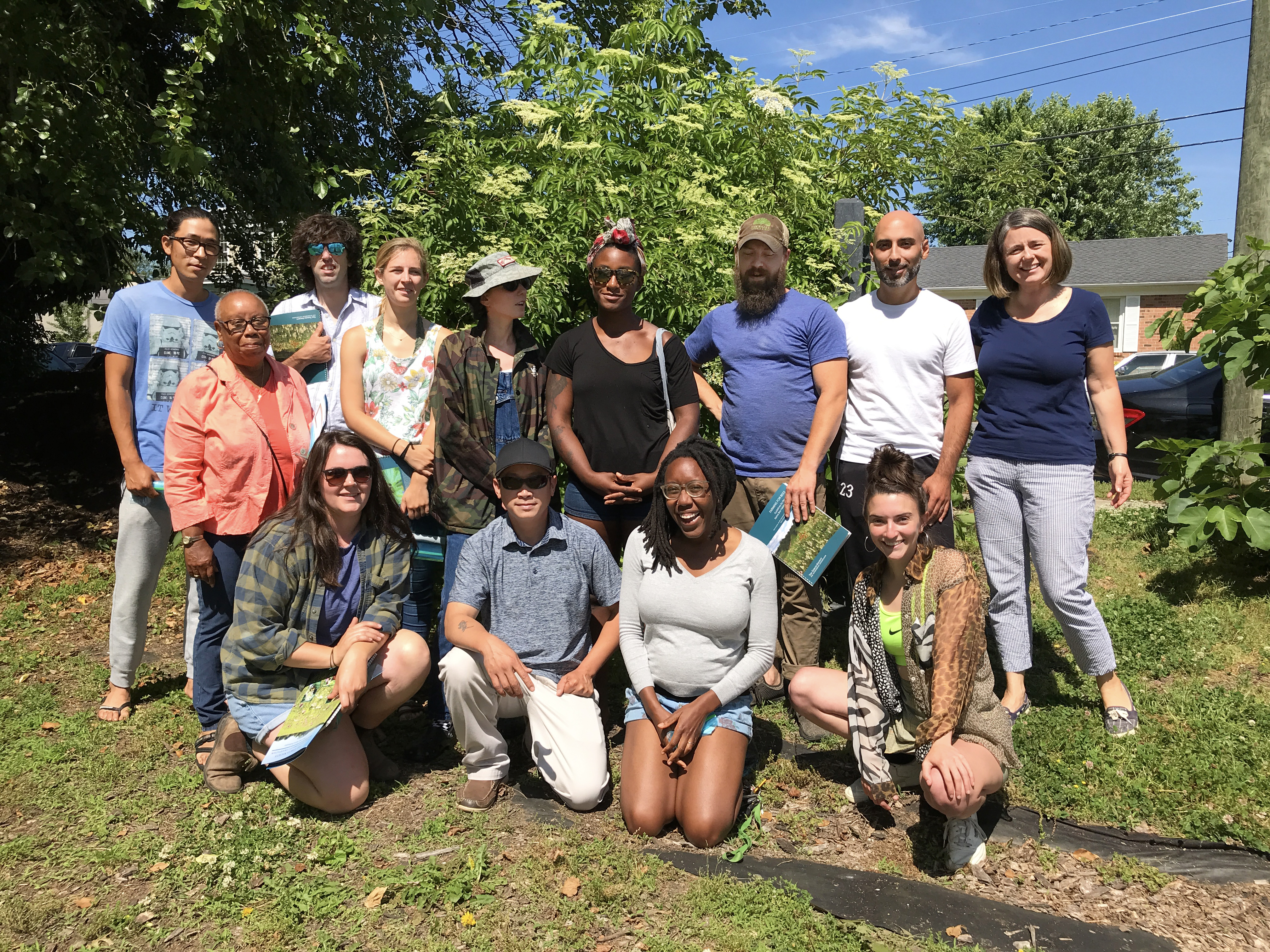 A group of smiling people gather in a lushly vegetated, urban setting.