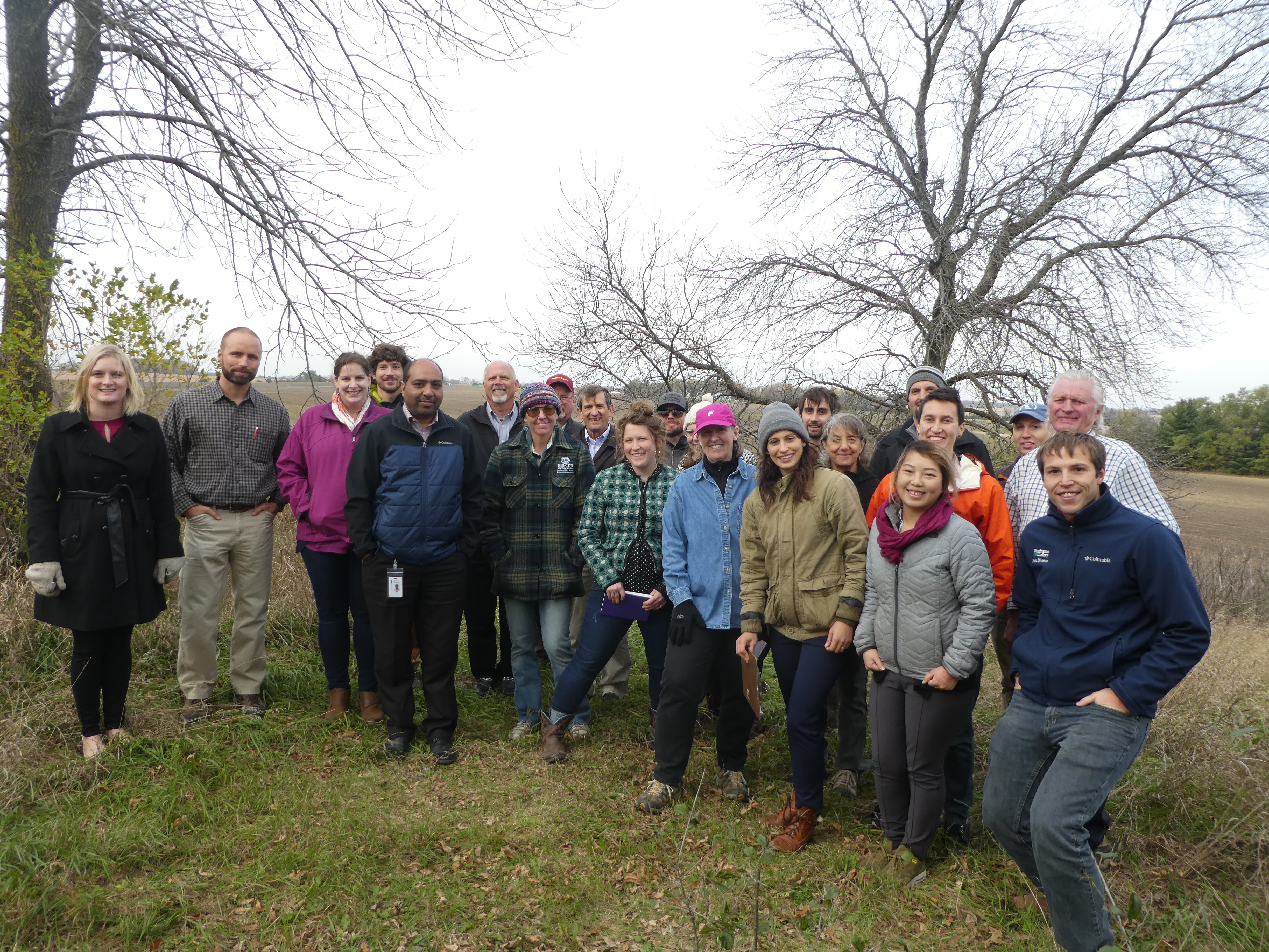 A group of people bundled in coats and hats smile for the camera as they gather in a wintry/late fall landscape.