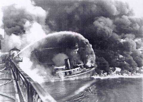 A firefighter sprays water from a bridge onto the burning Cuyahoga River in this historical photo, in black and white..