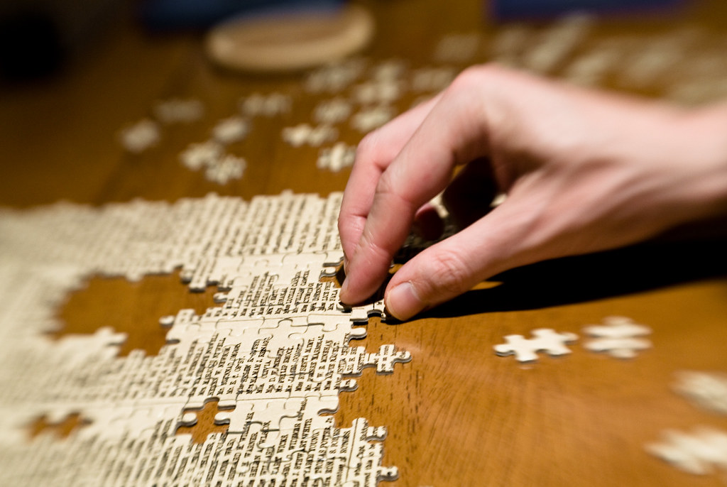 Pieces of a puzzle are being assembled on a dark, wooden table. A hand moves the pieces.