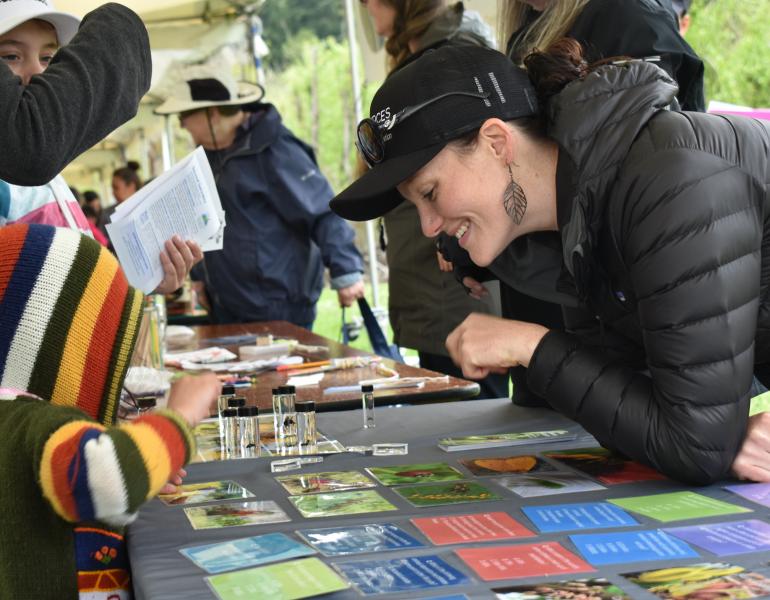 A smiling woman wearing a black coat and a black Xerces Society hat leans over a table. On the other side of the table, a kid with a colorful hoodie reaches for an assortment of small glass vials containing bee specimens. The woman and the kid are looking at the same vials and appear to be talking.