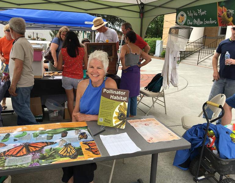  Phyllis Stiles, an older white woman with grey hair, presenting a table display at a local environmental festival. She is smiling and surrounded by information about pollinators. 