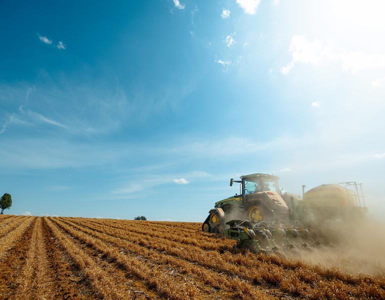 A tractor with a seed-planting attachment drives through a field planting corn seeds behind it as it travels.