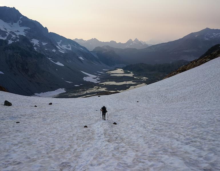 A Xerces conservation biologist hikes down from a small glacier that is now mostly just a snowfield. A chain of meltwater lakes can be seen in the distance, with smoke from nearby wildfires on the horizon.