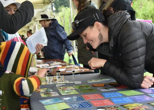A smiling woman wearing a black coat and a black Xerces Society hat leans over a table. On the other side of the table, a kid with a colorful hoodie reaches for an assortment of small glass vials containing bee specimens. The woman and the kid are looking at the same vials and appear to be talking.