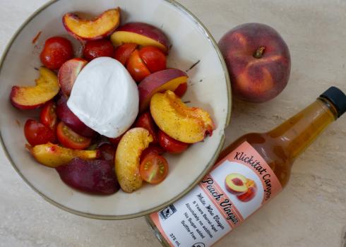 A bottle of vinegar lies on a table beside a bowl full of slices of peach and cherry tomatoes
