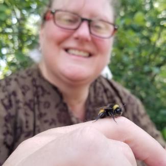 A woman smiles while looking at a fuzzy, black, and yellow bumble bee on her finger.