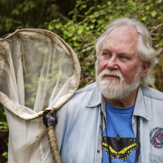Robert Michael Pyle, who founded the Xerces Society in 1971. He has grey hair and beard, and is wearing a blue denim shirt over a blue t-shirt with a black-and-yellow swallowtail butterfly design -- and is holding his butterfly net.