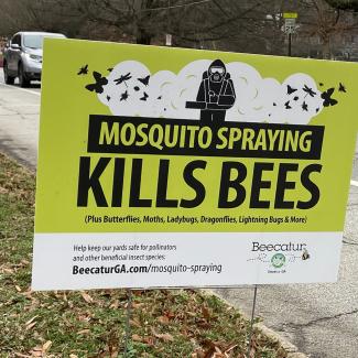 A roadside sign that is a bright green color with the message “MOSQUITO SPRAYING KILLS BEES” in black text. A silver-colored car and a bus drive past in the background.