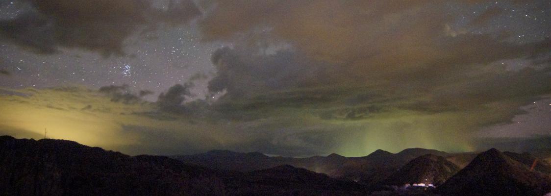 A mountainous scene is shown at night, with a small, glowing city in the lower right-hand corner. The mountains are dwarfed by an enormous sky, peppered with patchy clouds and swaths of stars. The sky has an unnatural glow (literally), with fields of yellow-green and orangish light, indicating light pollution.