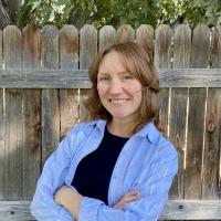 A woman with brown shoulder-length hair and a blue shirt smiles in front of a wooden fence.